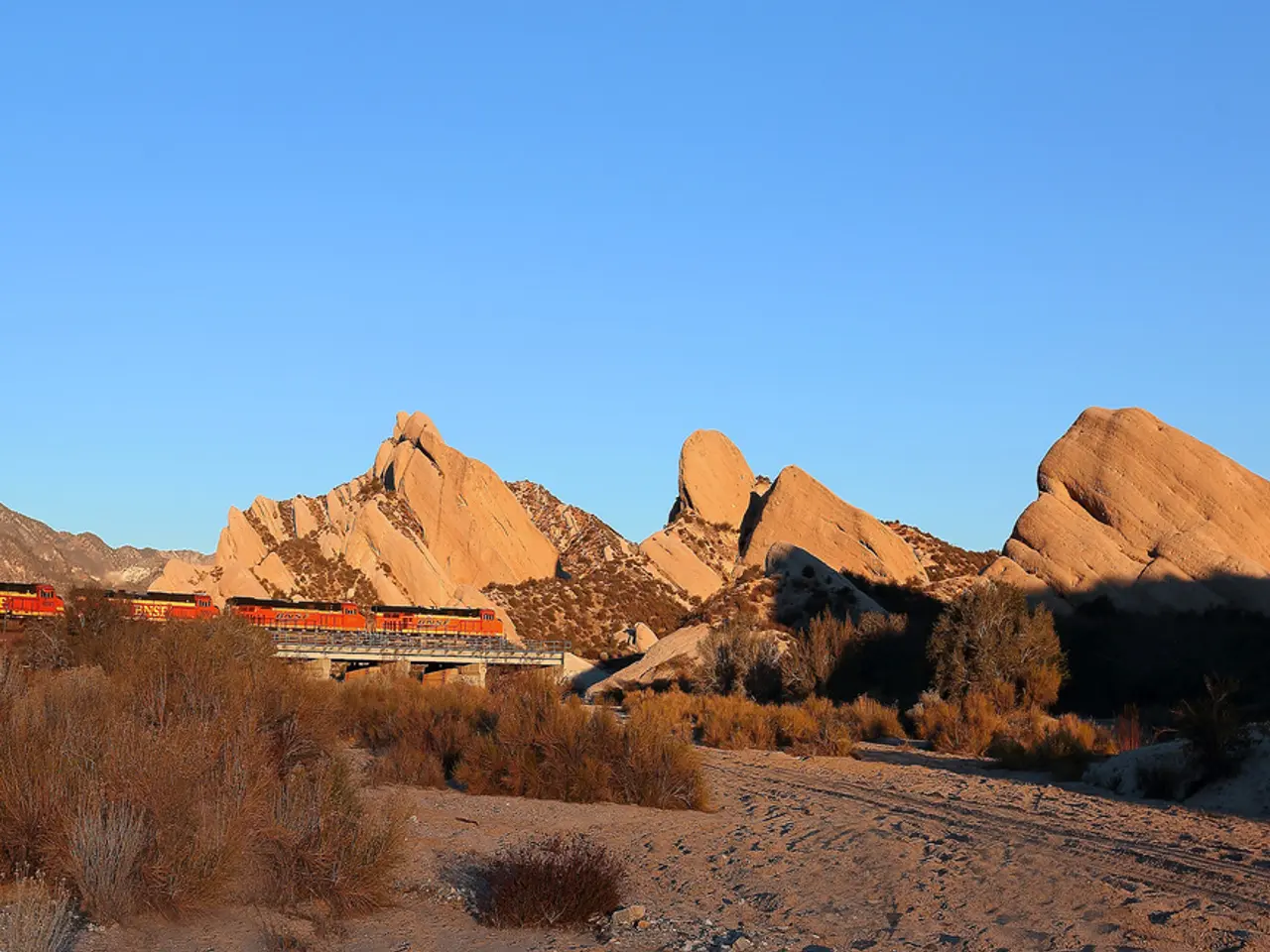 In this picture there is a train on the left side of the image, on the bridge and there are cliffs...