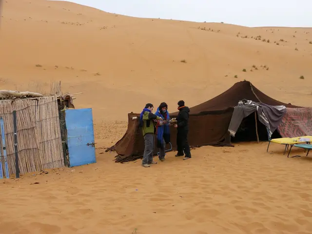 In this image we can see men standing on the sand, tents, thatched fences, shrubs, cots and sky.