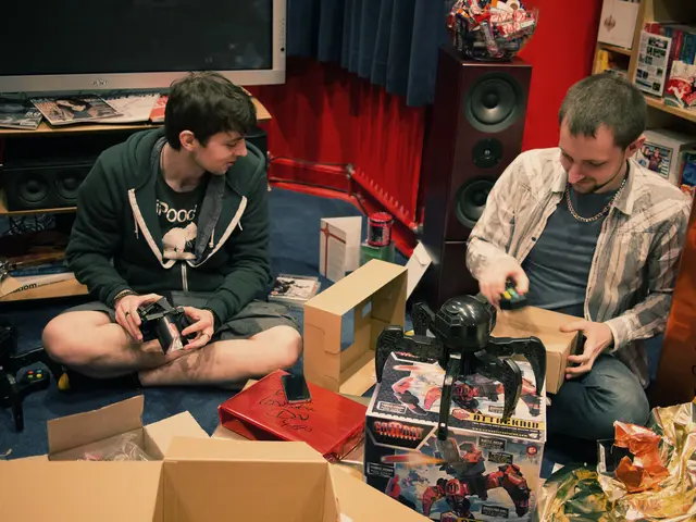 In this image, we can see two men sitting and there is a carton box, in the background there is a...